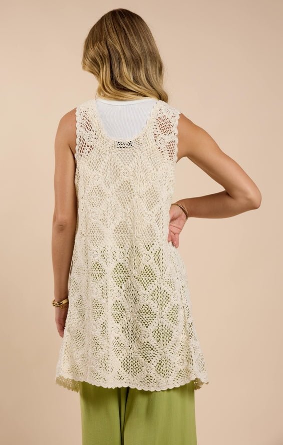 A woman faces away, wearing a small Fell For You Crochet Front Tie Cardigan by Sadie And Sage in cream over a white top and light green pants against a beige background—a chic addition to women’s clothing.