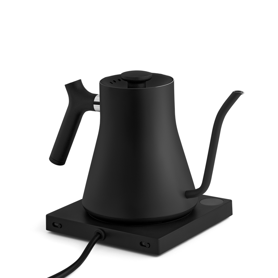 The Fellow Electric Kettle features a matte black finish, temperature control, a precision pour spout, and an ergonomic handle. It rests on a square base with an attached power cord.