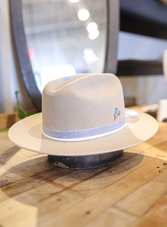 The Easy Drifter Felt Hat | Bluebonnet Champagne features a light beaver felt, blue ribbon, and hand-embroidered bluebonnet flowers, displayed on a wooden surface in front of a mirror.