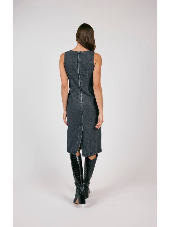 A woman faces away in the Fitted Denim Dress by Sage the Label—a sleeveless, knee-length black dress crafted from 100% cotton denim with a back slit—styled with black knee-high boots.