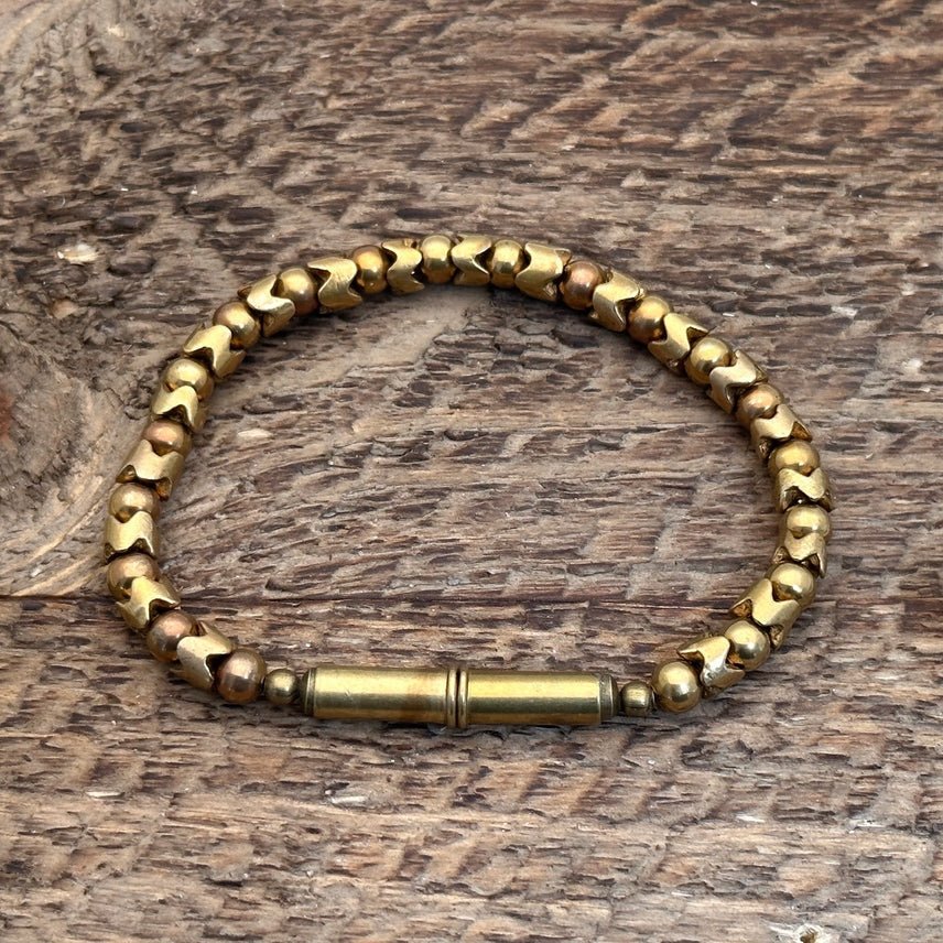 The Flint Beaded Single Bracelet by Tres Cuervos, featuring upcycled .22 shells and recycled glass beads with a cylindrical clasp, rests on a textured wooden surface.