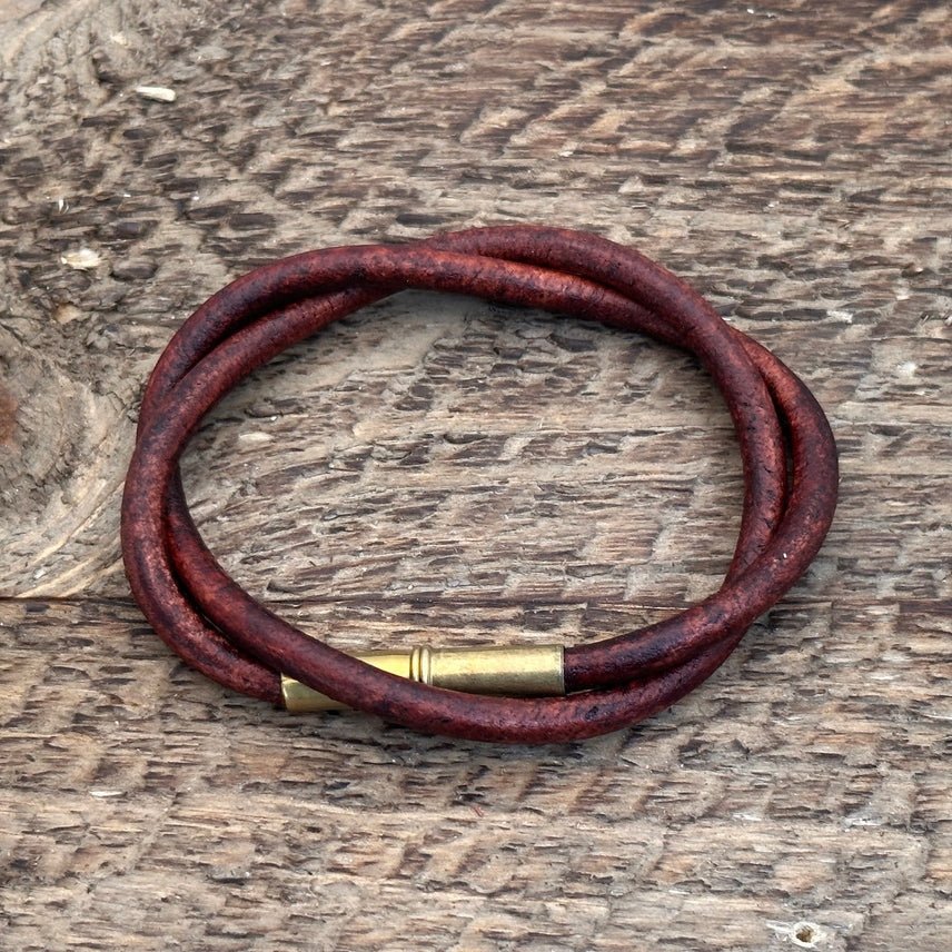 The Flint Bracelet by Tres Cuervos: a rustic brown leather cord accessory with a brass clasp, coiled into a circle and displayed on a textured wooden surface.