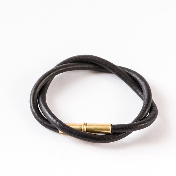 The Flint Bracelet by Tres Cuervos features black leather in a double wrap design with a gold cylindrical magnetic clasp—a modern twist on the classic style, shown on a white background.
