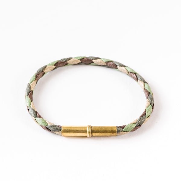 The Flint Single Waxed Canvas Bracelet by Tres Cuervos features green, beige, and brown braided strands with a cylindrical gold-colored magnetic clasp, shown against a white background.