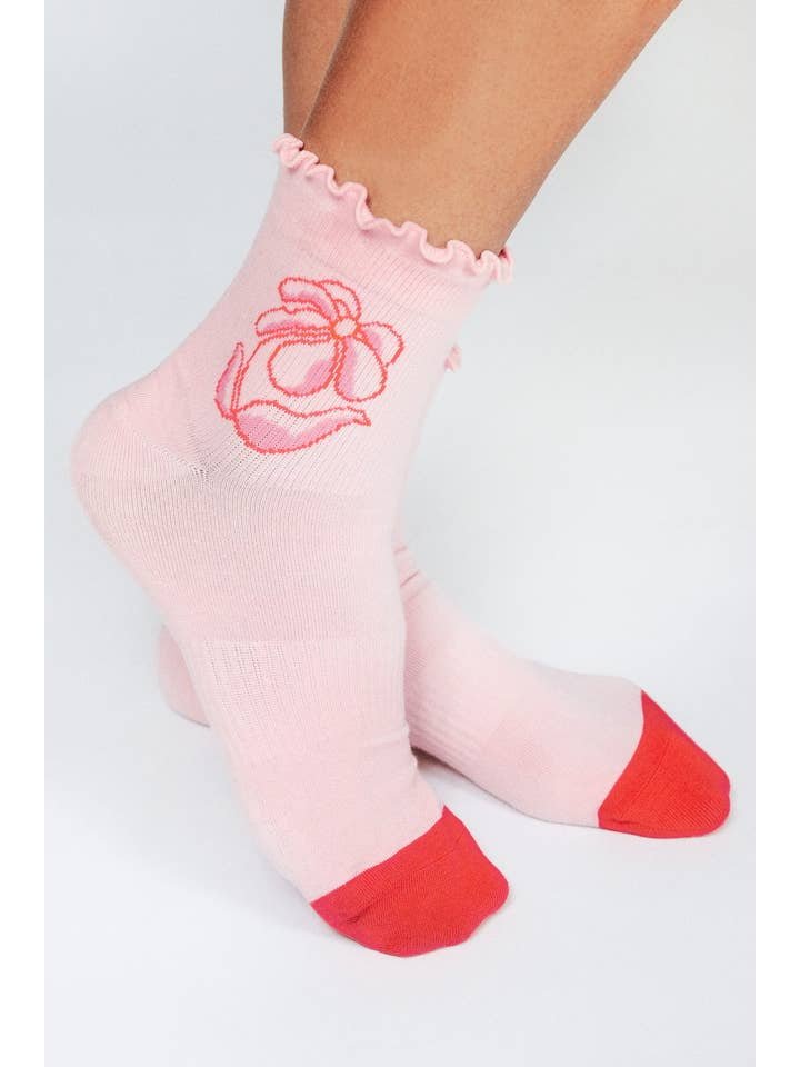 A pair of feet in Flora Ankle by Jordan Kay from Tailored Union—light pink socks with red toes, a floral side design, ruffled edge—crafted from premium combed cotton for comfort.