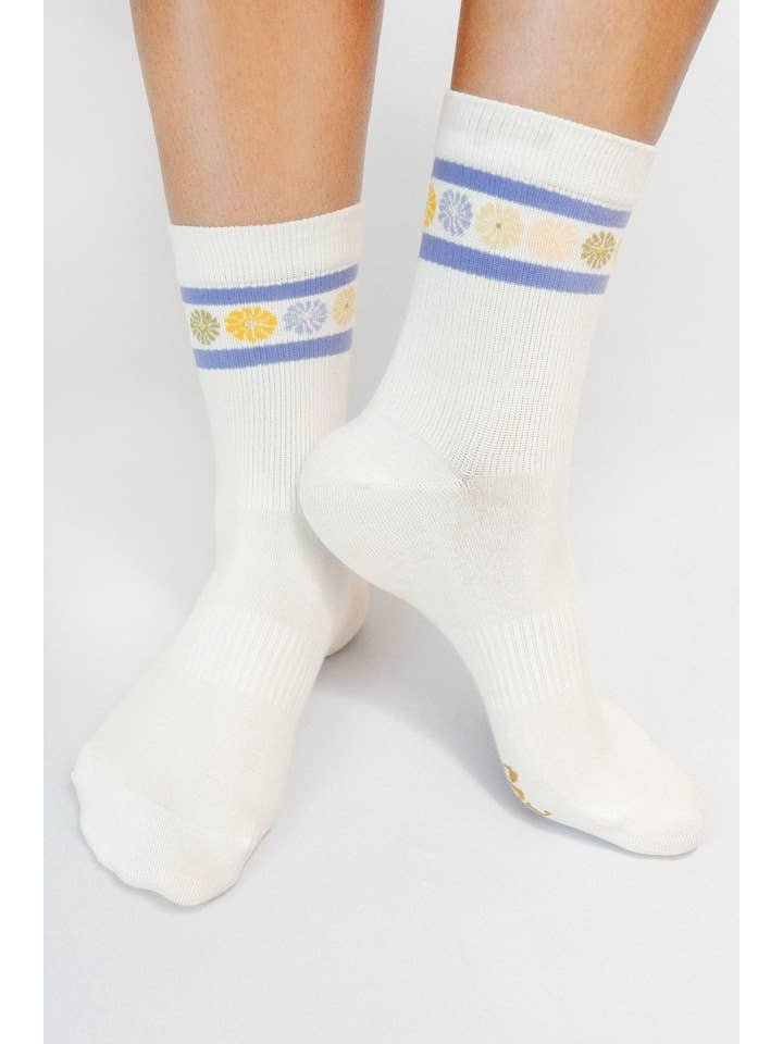 A pair of feet in Tailored Union's Flower Crown Knit Ankle Sock—white with blue stripes and yellow and purple flower accents near the top.