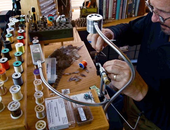 At a workbench, an angler crafts hand tied flies using the Guide Curated Fly Set by Mollyjogger—ideal for trout fishing and passionate fly enthusiasts, surrounded by threads, materials, and essential tools.