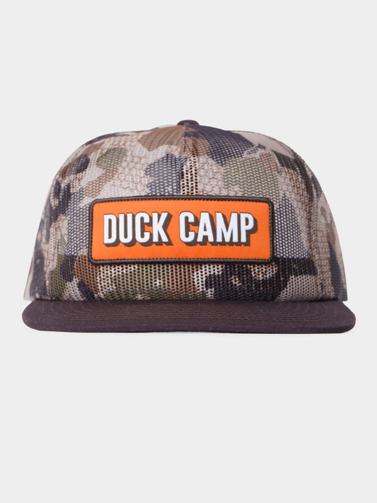 Duck Camp’s Gulf Trucker in Wetland features a camouflage design, flat brim, breathable mesh panels, and an orange front patch with "DUCK CAMP" in white letters.
