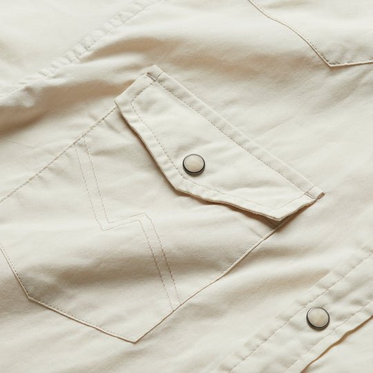 Close-up of a H Bar B Snapshirt | Riverbed Oxford by Howler Brothers, showing the beige pocket with flap and pearl snaps, detailed stitching, and part of another western shirt pocket in the background.