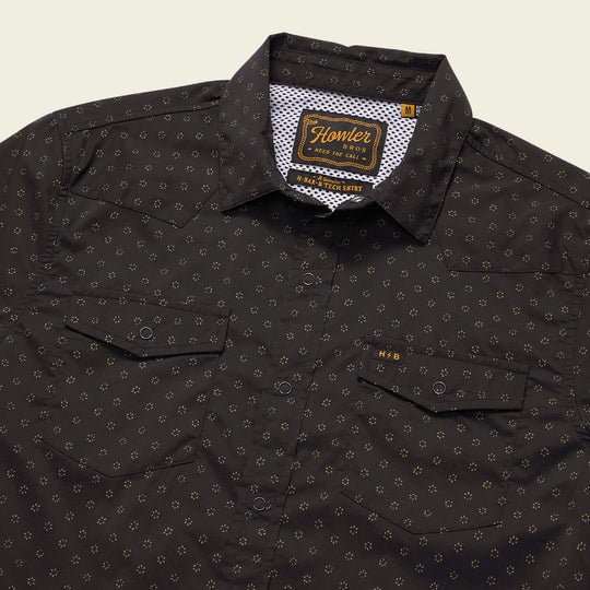 The Howler Brothers H Bar Tech Long Sleeve in Little Puddles features a black lightweight stretch fabric with a small gold geometric pattern, two chest pockets, a white patterned inner collar, and a vented back yoke for comfort.