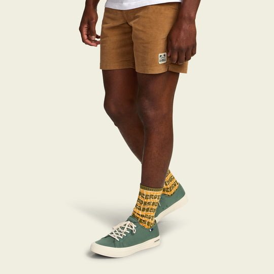 A person wearing Howler Brothers' HB Flow shorts, green sneakers with white soles, and vintage-inspired athletic crew socks stands against a plain light background.