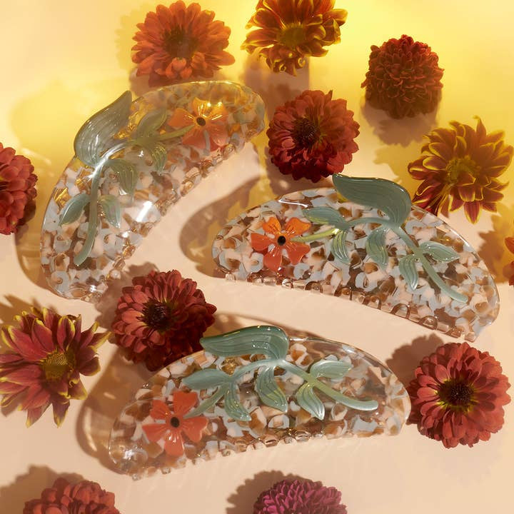The Hesperides Large Hair Claw by Winona Irene is displayed among red and orange chrysanthemum flowers on a pastel yellow background, highlighting this charming accessory.