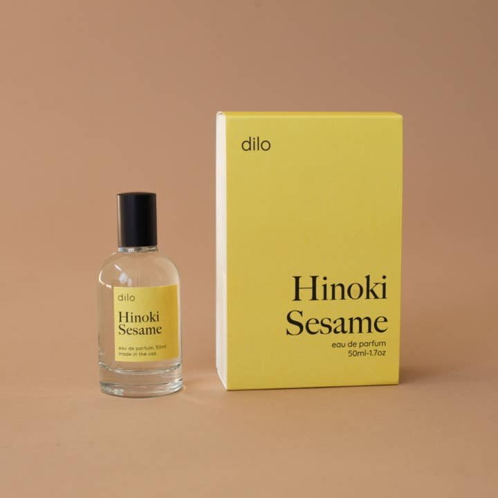A 50ml bottle and box of dilo's Hinoki Sesame Eau De Parfum with yellow labels rest on a brown background—this inviting scent blends sandalwood and hinoki sesame notes.