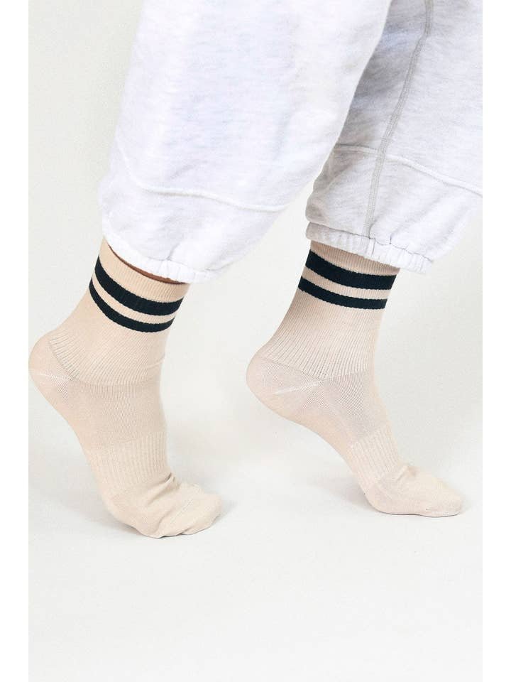 Someone stands on tiptoes against a plain background, wearing the Jouer Ankle Sock-Oat by Tailored Union—with two black stripes—and light gray sweatpants.