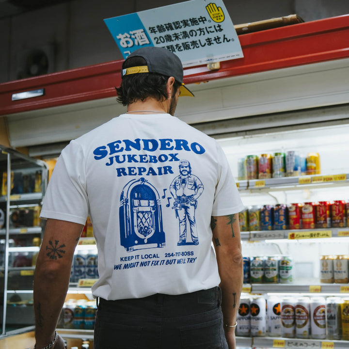A man wears the Sendero Provisions Co. Jukebox Repair T-Shirt—a white cotton-poly tee with a soft-touch "Sendero Jukebox Repair" print—while standing by drink shelves in a convenience store.