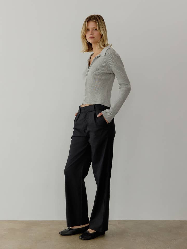 A woman stands against a plain wall wearing the light gray Kieran Top by All Row—a collared zip-up knit with ribbed detailing—paired with black wide-leg trousers and black shoes, her hands in her pockets.