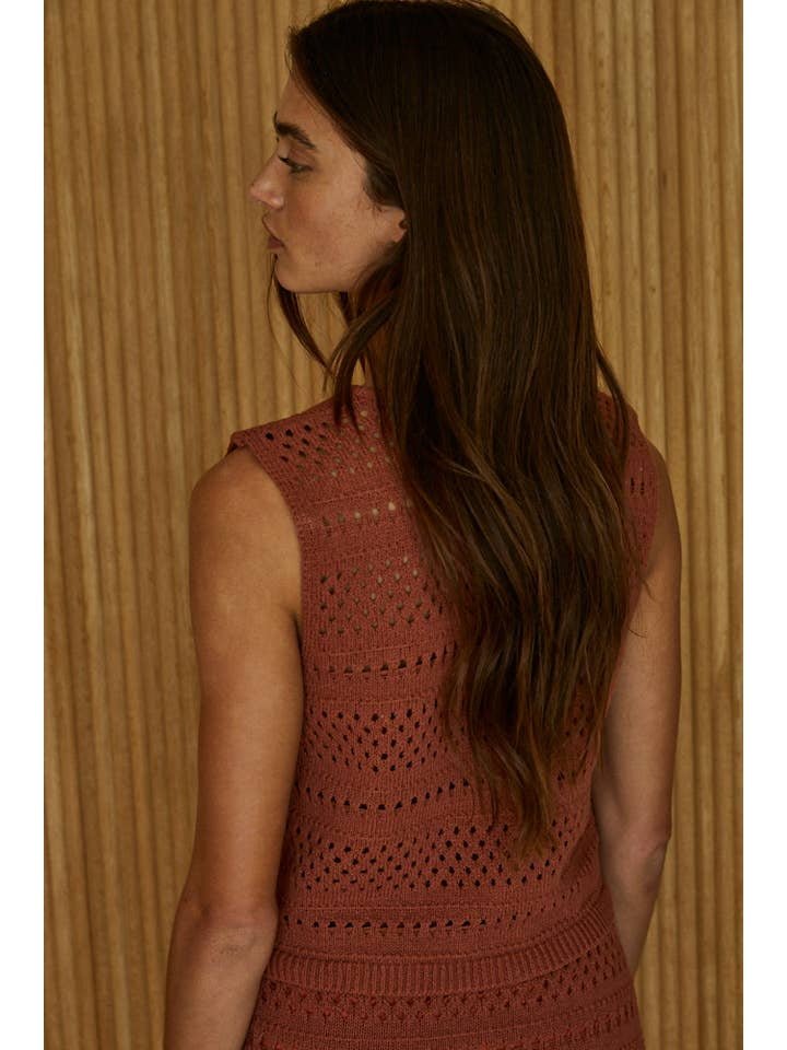 A woman with long brown hair, wearing the By Together Knit Sweater Cotton Crochet Best Top, stands facing away in front of a light wooden panel wall.