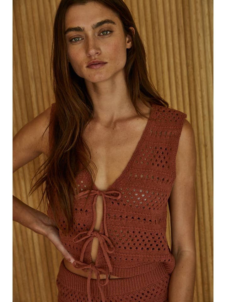 A woman with long brown hair models the By Together Knit Sweater Cotton Crochet Best Top, a sleeveless rust vest with tie closures, in front of a wooden background for a boho-chic look.