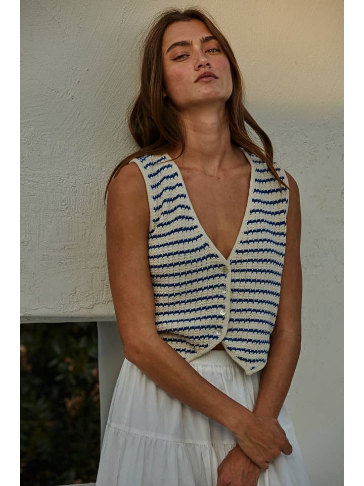 Woman wears the By Together Knit Sweater Cotton Striped Buttondown V-Neck Vest in cream and blue over a white dress, standing against a textured white wall.
