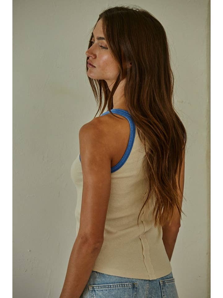 A woman with long brown hair wears the By Together Knit Tank Top, a sleeveless beige knit style with blue trim, paired with light blue jeans, as she stands slightly turned from the camera against a plain wall.
