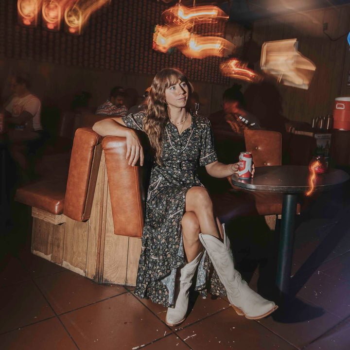 A woman in the La Dahlia Dress by Sendero Provisions Co. and white boots sits on a booth seat in a dimly lit bar, holding a can of beer, with blurred lights and people in the background.