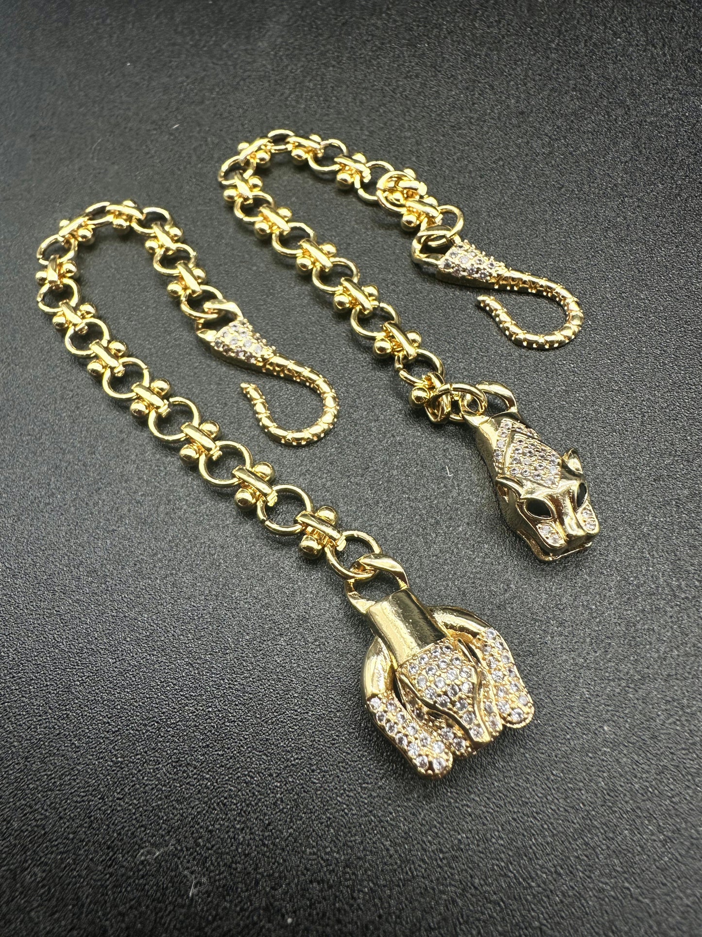 Two La Prise Bracelets by Minh Atelier, crafted in an 18k gold-plated finish with diamond-studded panther head clasps and decorative hooks, are displayed on a black surface.