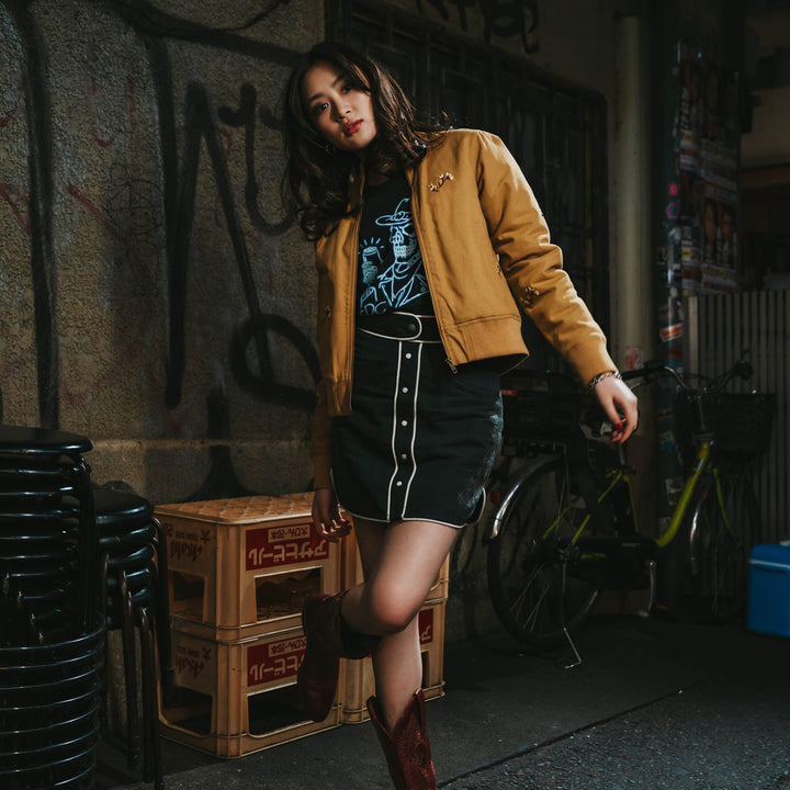 A woman wearing the La Tierra Jacket by Sendero Provisions Co., a graphic tee, black skirt, and red boots poses in an urban alleyway beside stacked crates and a bicycle.