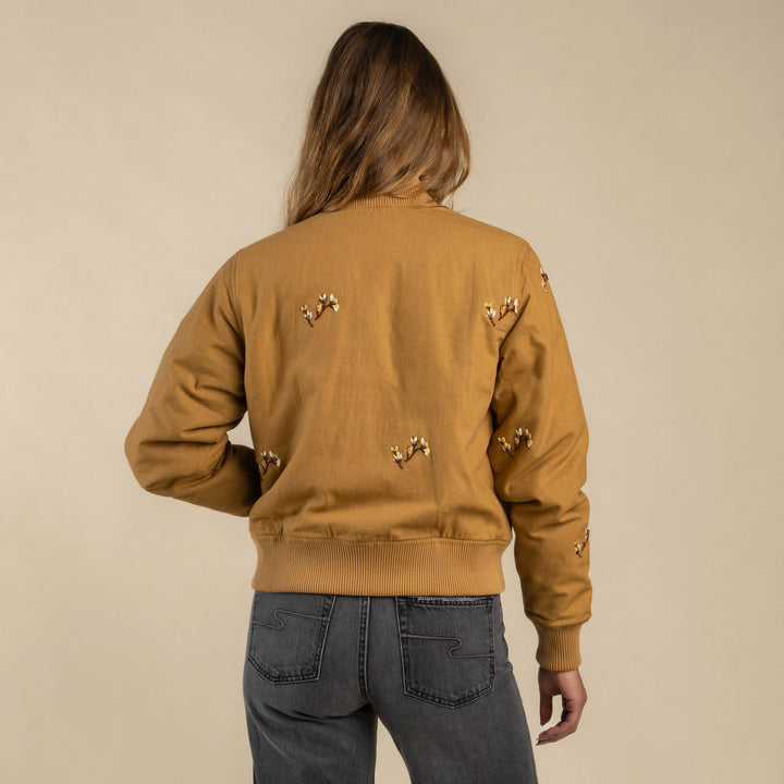 A person with long hair wears the Sendero Provisions Co. La Tierra Jacket and gray jeans, standing with their back to the camera against a beige background.