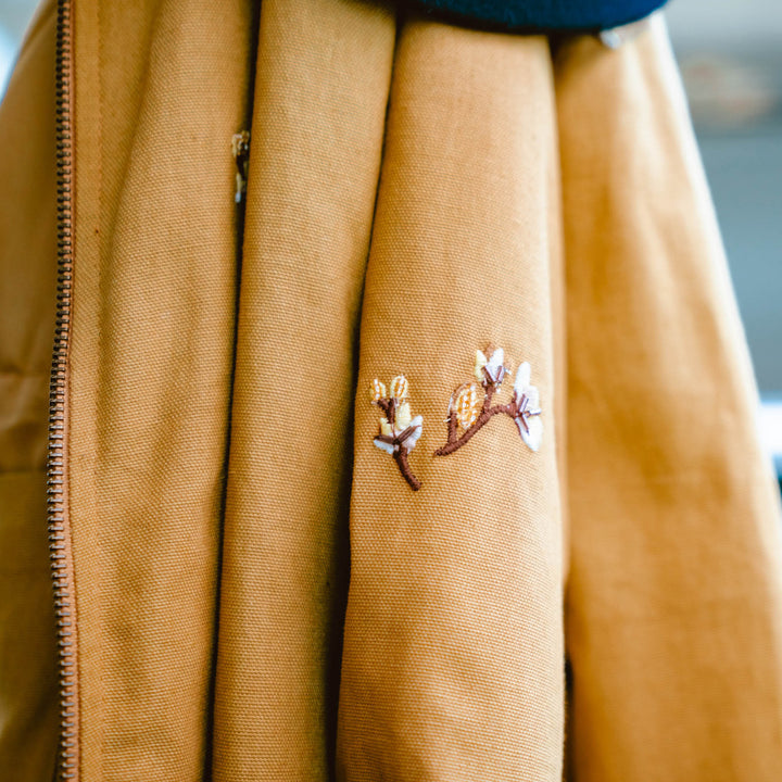 Close-up of the La Tierra Jacket by Sendero Provisions Co.—a cotton canvas jacket with a left-side zipper and delicate white and pink floral embroidery on the right, highlighting detailed embroidered outerwear.