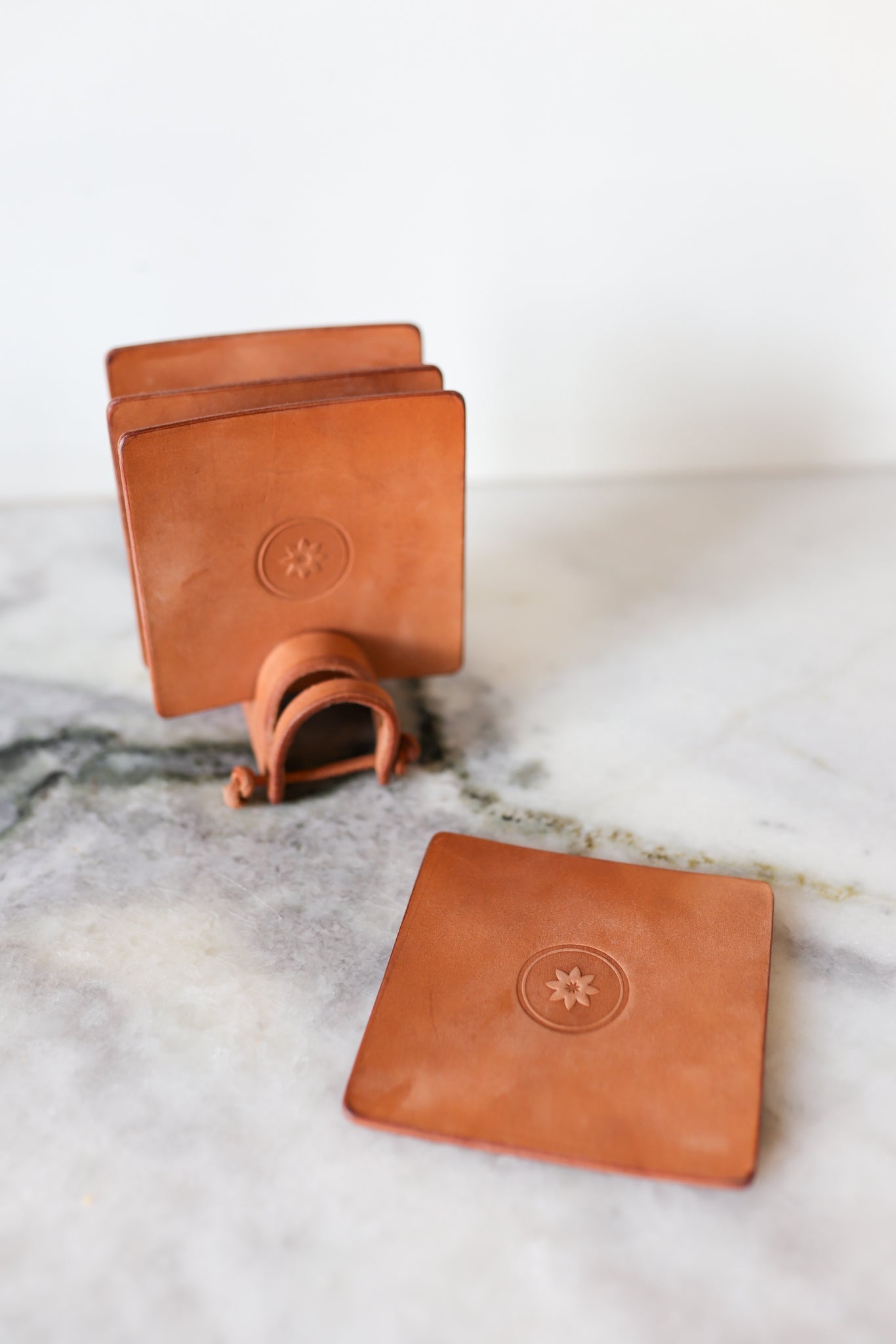 The Ballad of the Bird Dog Leather Coaster 4 Piece Set features round coasters with a star design, shown on marble with one displayed in front and three stacked in the handmade leather stand, perfect for desk protection.