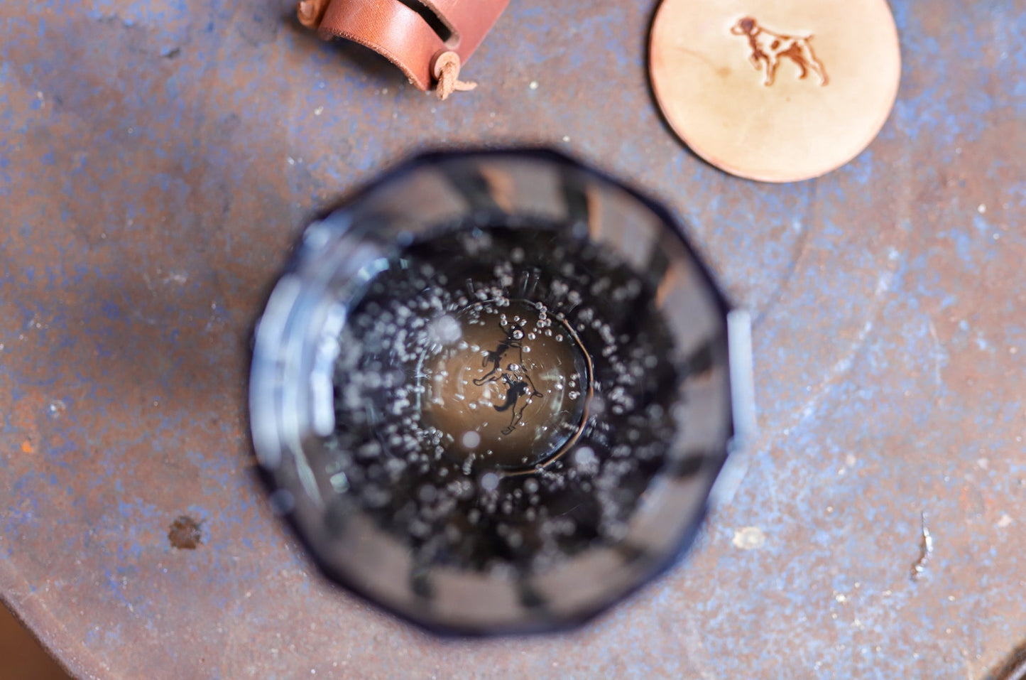 Top-down view of a glass with fizzy, dark liquid next to the Ballad of the Bird Dog Leather Coaster 4 Piece Set—handmade coasters embossed with a dog, perfect for stylish desk protection.