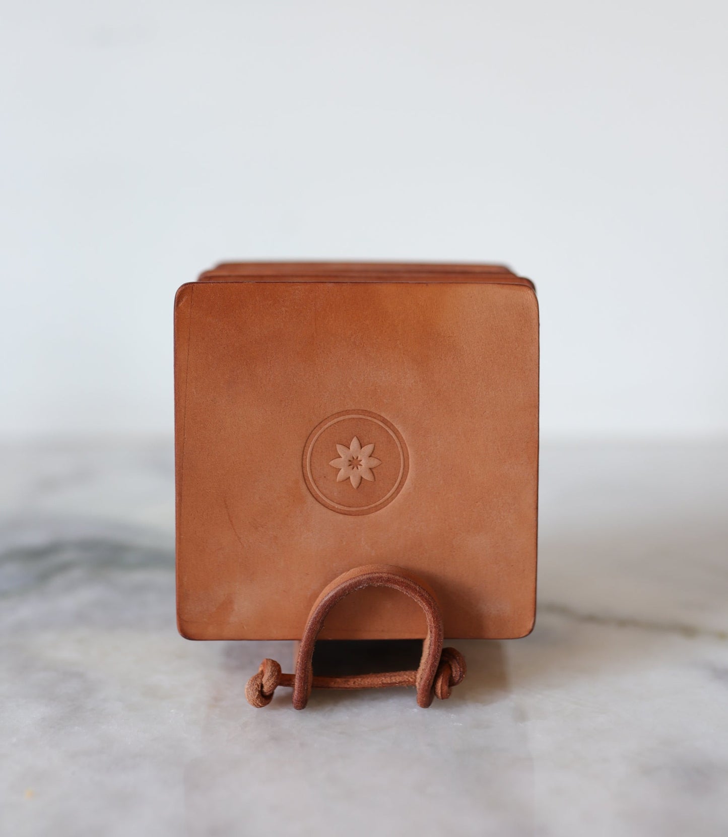 The Leather Coaster 4 Piece Set by Ballad of the Bird Dog sits on a marble surface in a brown handmade leather box with a flower emblem and bottom strap, perfect for protecting your desk or storing round coasters.