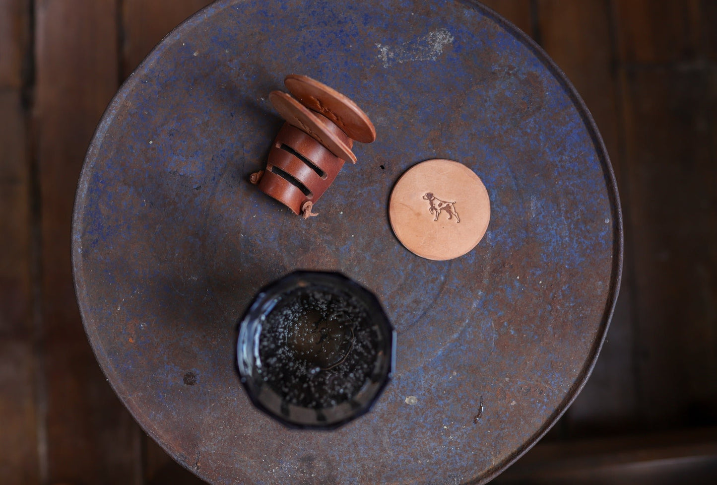 A round metal table with dark glass, a brown ceramic cup on its side, and Ballad of the Bird Dog’s Leather Coaster 4 Piece Set—handmade round leather coasters with dog engraving for stylish desk protection.