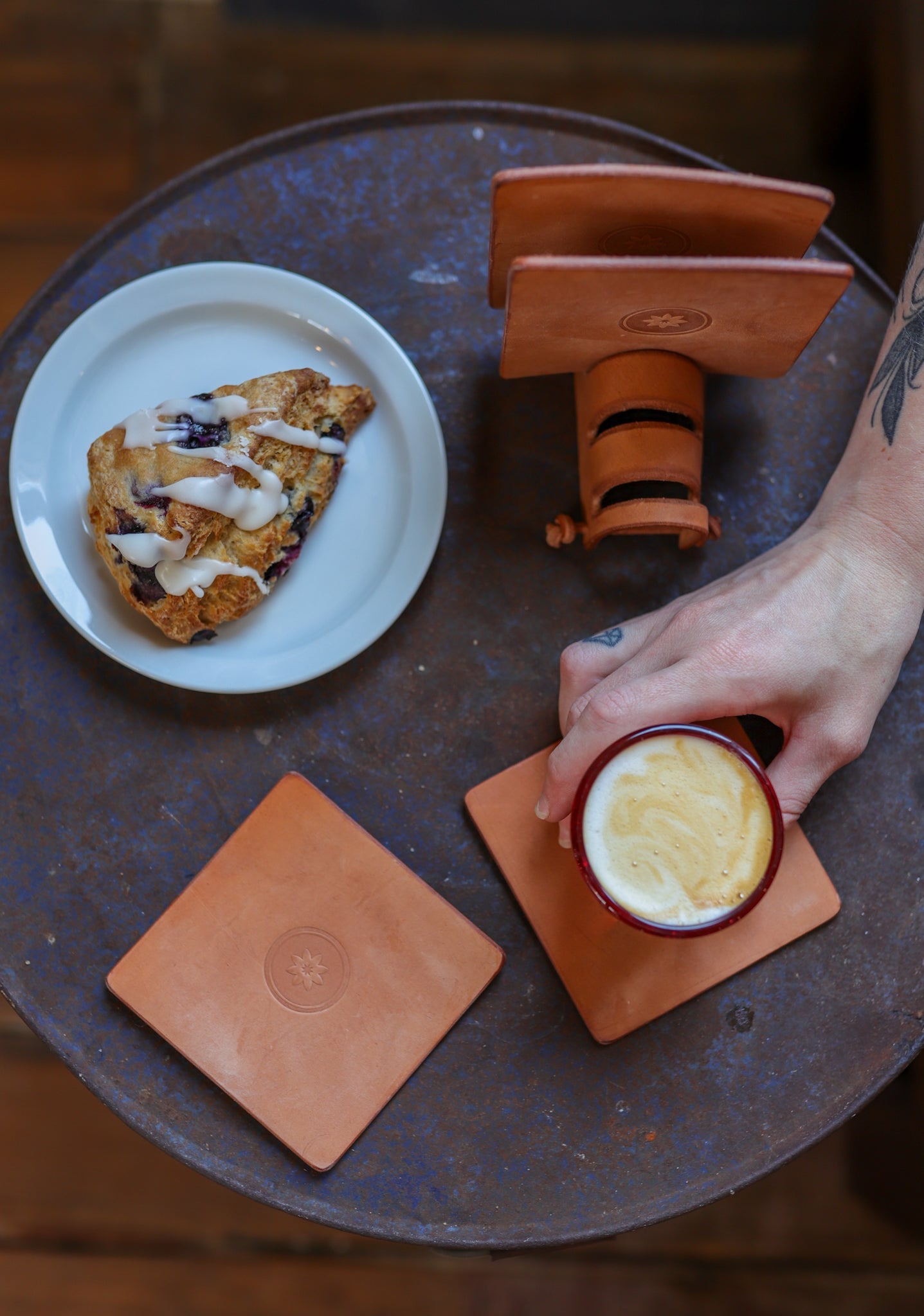 A hand holds a cup of coffee by a pastry on a round table, with the Leather Coaster 4 Piece Set from Ballad of the Bird Dog protecting the desk and showcasing your style.