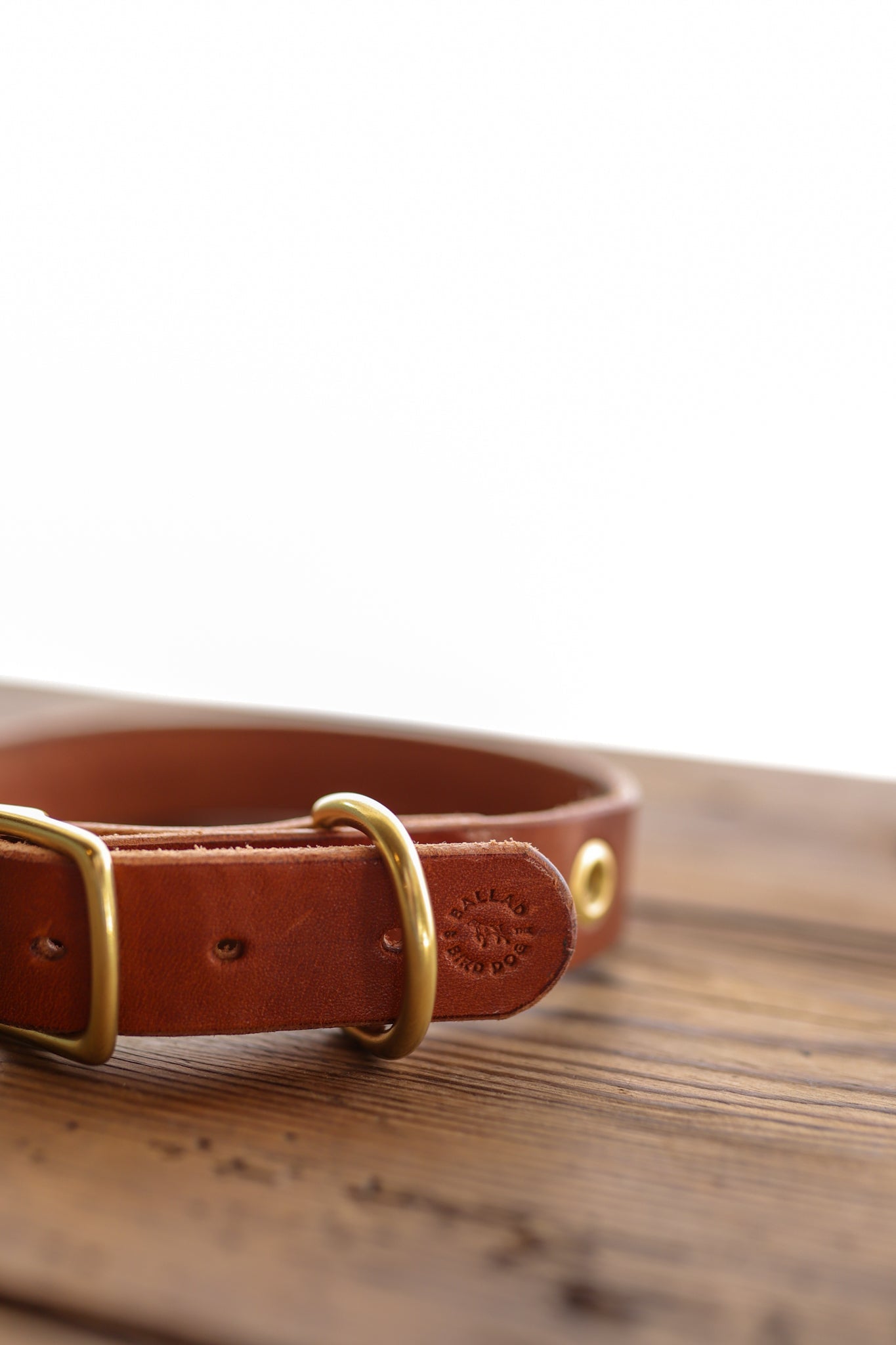 The Leather Dog Collar by Ballad of the Bird Dog, crafted from brown English bridle leather with brass hardware, is displayed on a wooden surface against a white background.