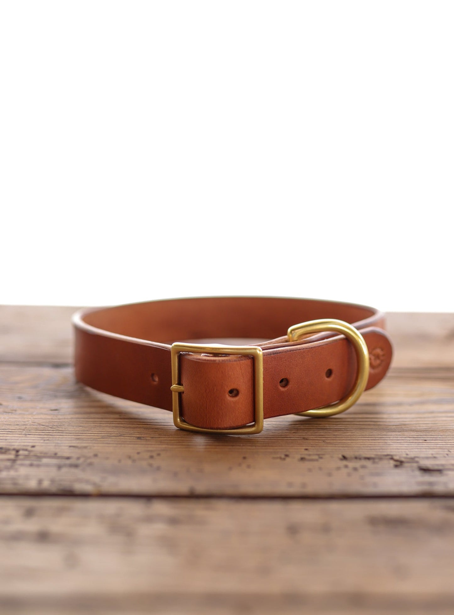 The Leather Dog Collar by Ballad of the Bird Dog, a handmade brown English bridle leather collar with a gold buckle and D-ring, is shown on a wooden surface against a white background.
