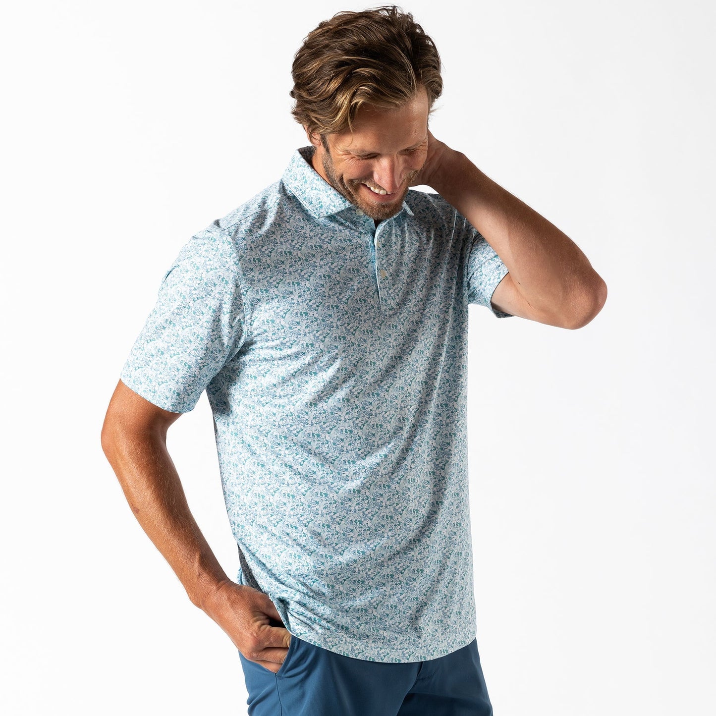 A man in a light blue patterned Duck Head Long Drive Performance Polo and blue pants stands against a white background, looking down with one hand behind his neck.