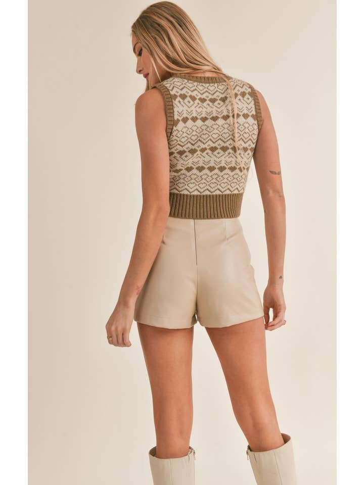 A woman stands facing away against a neutral background, wearing the Lovers Vest by Sage the Label—a sleeveless patterned sweater vest—paired with beige high-waisted shorts and knee-high boots.
