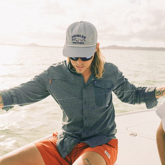 A man in a grey Howler Brothers cap, sunglasses, orange shorts, and a Lotsa Dots Matagorda Long Sleeve shirt by Howler Brothers with UPF 35 sits on a boat with water and land in the background.