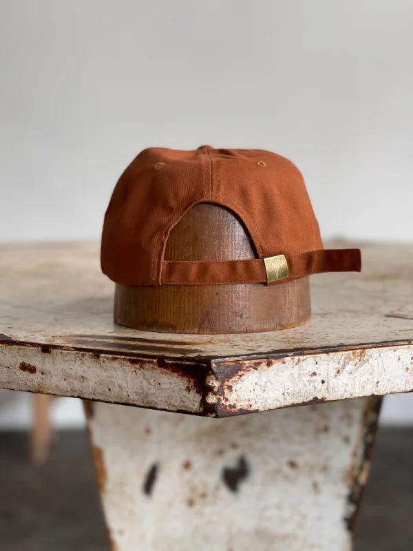 The Mescalito Field Hat by Ballad of the Bird Dog—a brown cotton cap with an adjustable strap—rests reversed on a rustic table, ready for your next trip to Mescalito Coffee.