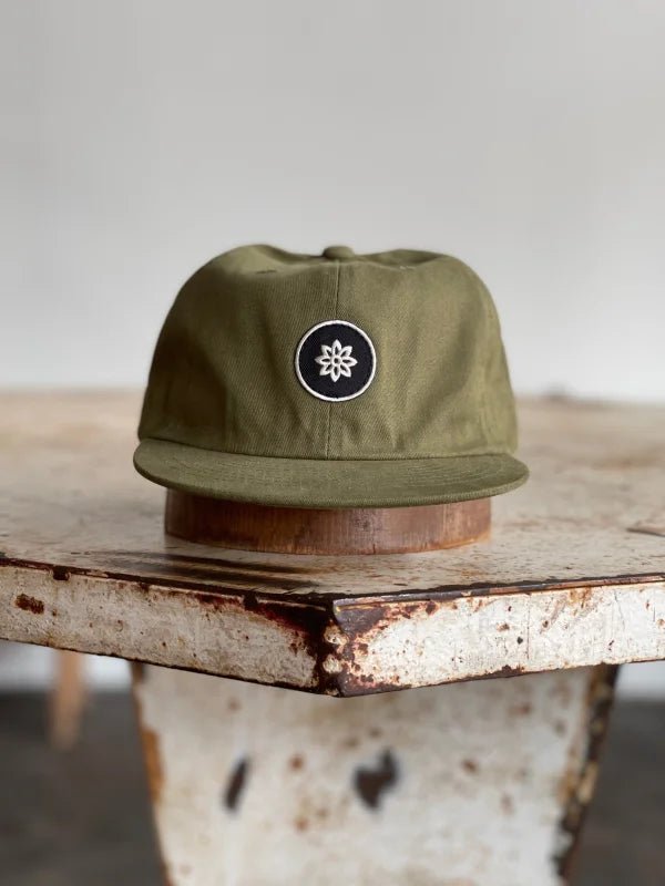 The Mescalito Field Hat by Ballad of the Bird Dog features an olive green cotton cap with a black and white embroidered flower logo—ideal for coffee shop style or mornings at Mescalito Coffee.