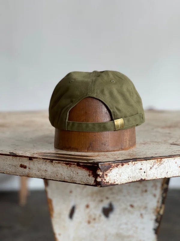 The Mescalito Field Hat by Ballad of the Bird Dog, in olive green cotton with an adjustable strap, rests on rustic wood—an ideal accessory for your next stop at Mescalito Coffee.