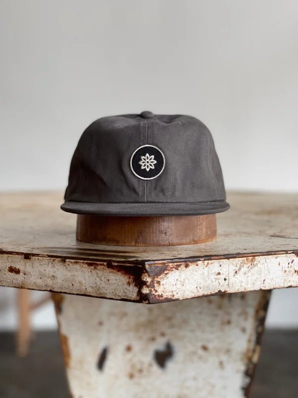 The Mescalito Field Hat by Ballad of the Bird Dog—a dark gray cotton cap with a white flower emblem—rests on a weathered metal table, ready for your next trip to Mescalito Coffee.