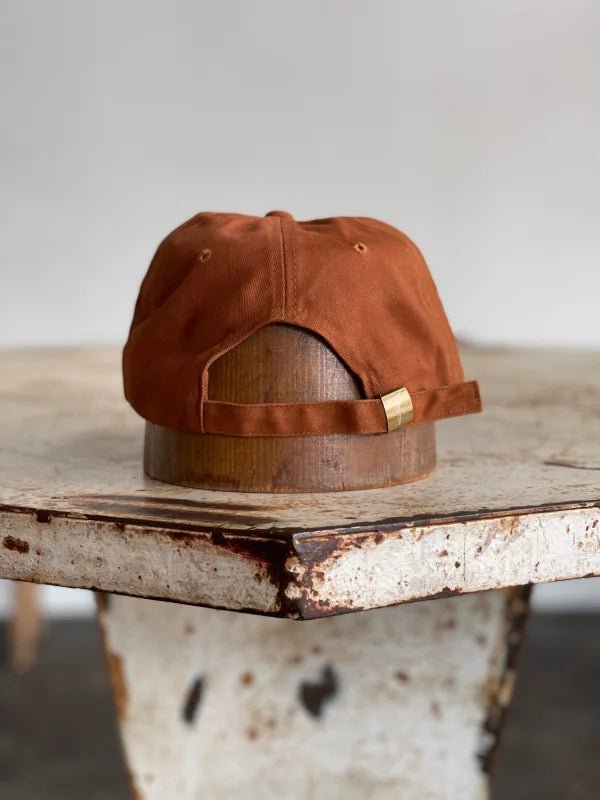 The Mescalito Field Hat by Ballad of the Bird Dog in coffee brown is displayed from the back on a wooden stand atop a rustic, weathered table.