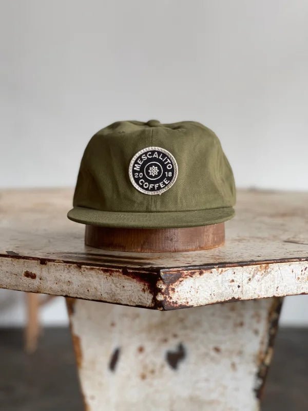 The Mescalito Field Hat by Ballad of the Bird Dog, featuring a circular "Mescalito Coffee" patch, rests on a worn rustic wooden table.