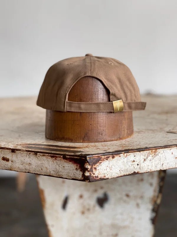The Mescalito Field Hat by Ballad of the Bird Dog, in brown cotton, is displayed rear-view on a cylindrical wooden stand atop a worn white table.