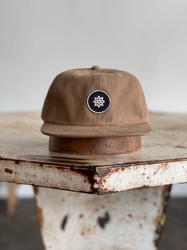 The Mescalito Field Hat by Ballad of the Bird Dog, made from brown cotton and featuring a black-and-white geometric flower logo, is perfect for Mescalito Coffee fans.