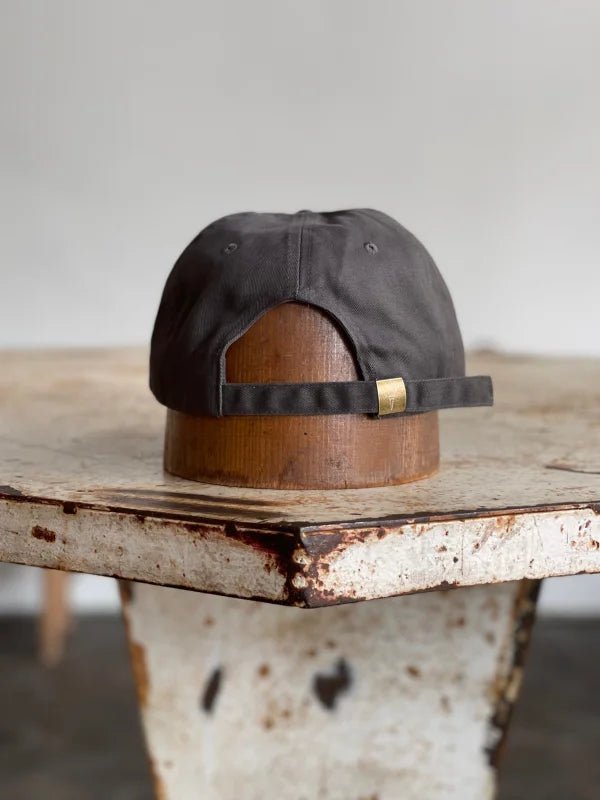 The Mescalito Field Hat by Ballad of the Bird Dog is shown from the back, resting on a wooden stand atop a rustic table—an ideal pick for fans of Mescalito Coffee or anyone searching for the perfect coffee shop hat.