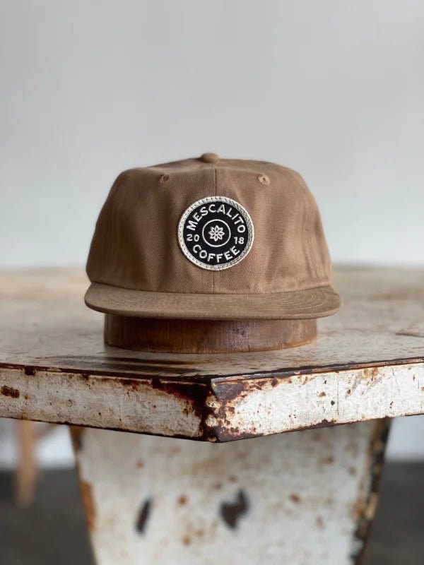 The Mescalito Field Hat by Ballad of the Bird Dog features a brown cotton design with a black and white "Mescalito Coffee" patch—an ideal addition to any coffee shop hat collection.