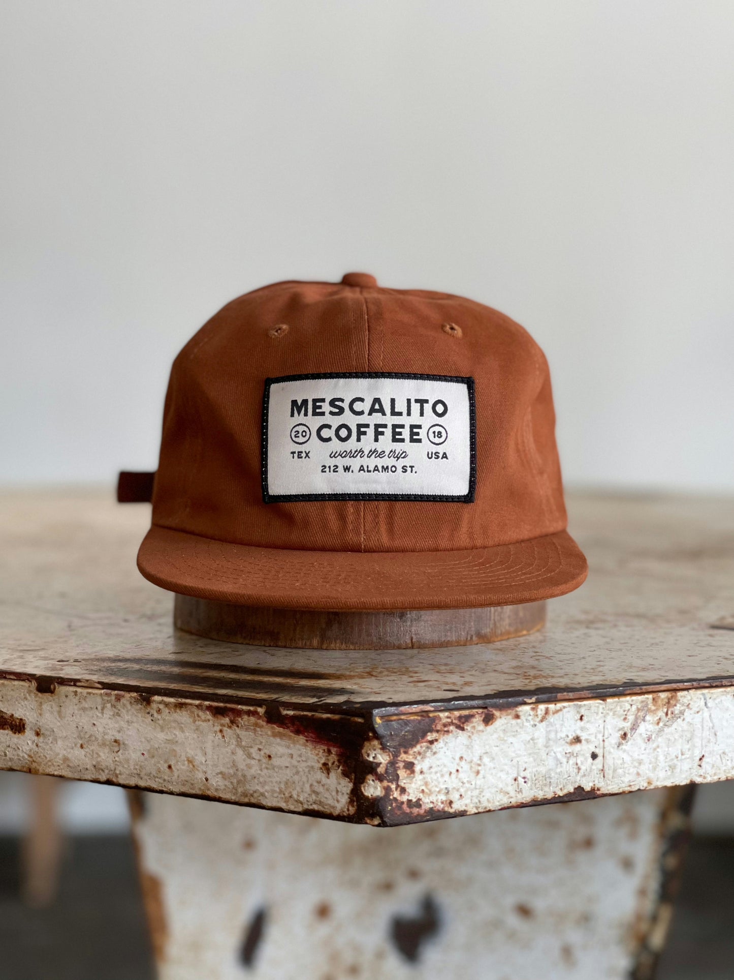 The Mescalito Field Hat by Ballad of the Bird Dog, featuring a "Mescalito Coffee" patch and crafted from brown cotton, rests on a rustic table—ideal for any coffee shop hat collection.
